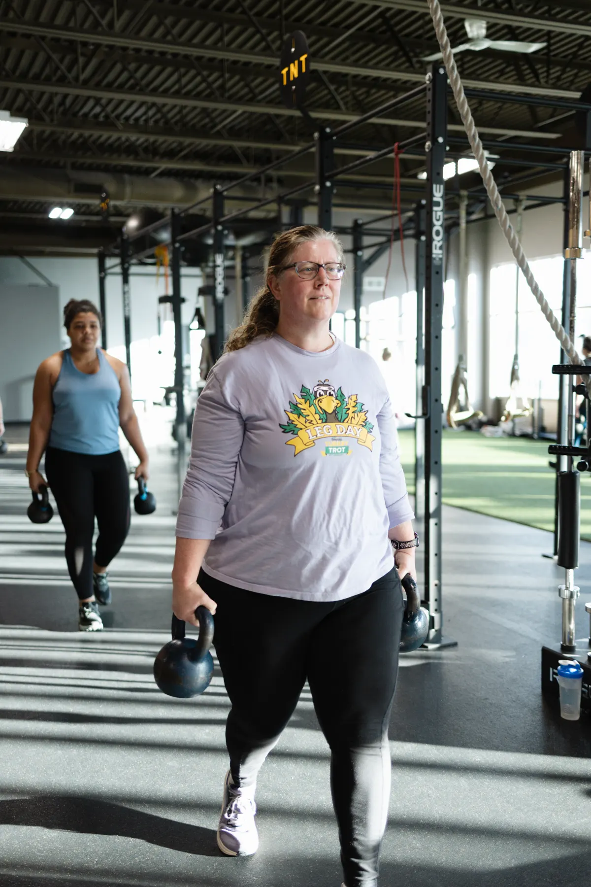 Adult member carrying heavy dumbbells across the TNT Fitness training floor while a coach watches